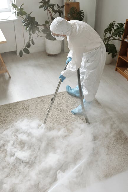 A professional cleaner in full protective white suit, face mask, and gloves is performing deep cleaning on a light-colored carpet in a modern living room. The cleaner is using a portable steam cleanin